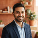 Portrait headshot of a smiling South Asian man in a modern, professional setting with warm terracotta and white decor.